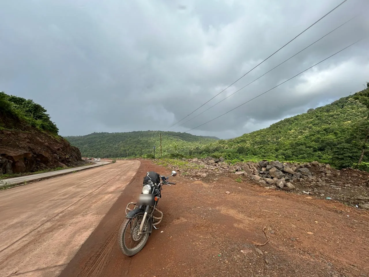 A Royal Enfield Bullet 500 parked on a red-earth ghat road under monsoon clouds